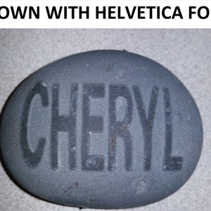 May include: A grey stone with the name "CHERYL" written in black Helvetica font.