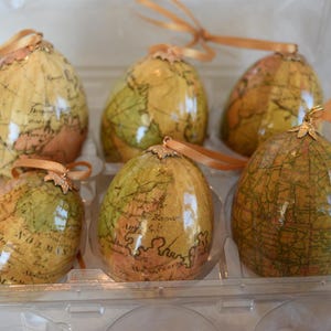 May include: Six globe-shaped ornaments with a vintage map design. The ornaments are in a clear plastic container. Each ornament has a gold-colored cap and a gold ribbon for hanging. The map design features shades of tan, green, and brown.