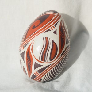 May include: A white ceramic egg with a geometric design in shades of orange and brown. The egg is decorated with a pattern of lines, swirls, and dots.