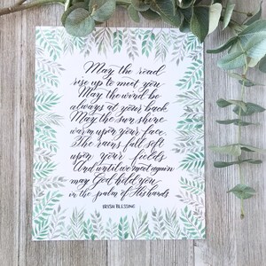 May include: A framed print with an Irish blessing in elegant calligraphy. The text, in black script, is surrounded by a watercolor border of green leaves. The words include "May the road rise up to meet you" and "Irish Blessing."