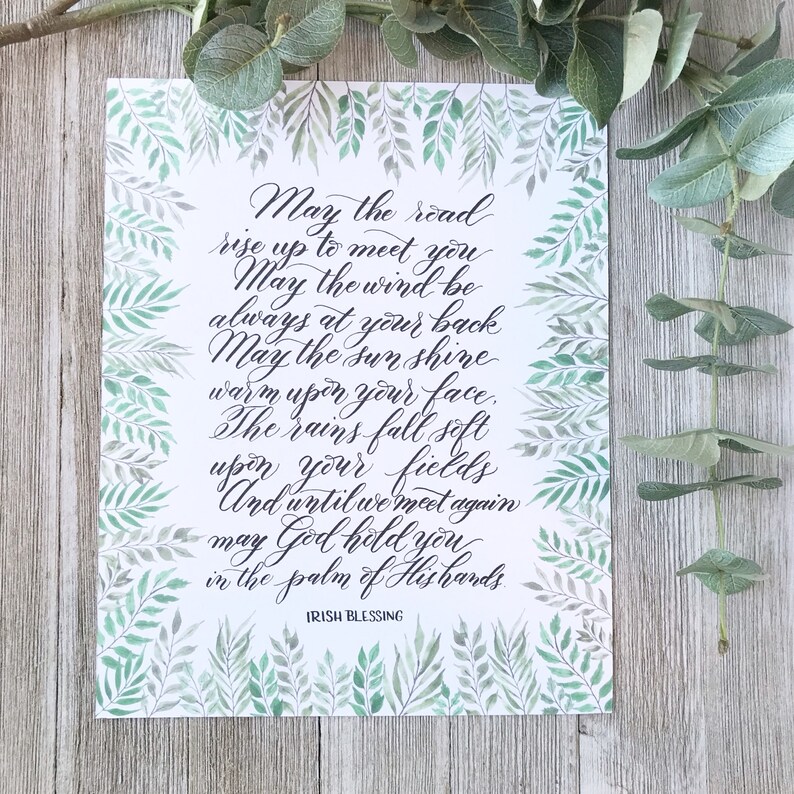 May include: A framed print with an Irish blessing in elegant calligraphy. The text, in black script, is surrounded by a watercolor border of green leaves. The words include "May the road rise up to meet you" and "Irish Blessing."