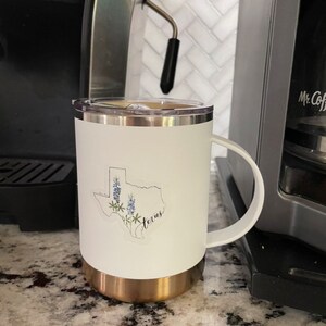 May include: White travel mug with a gold base and a handle. The mug features a sticker of the Texas state outline with bluebonnet flowers and the word "Texas". The mug is next to a coffee machine.