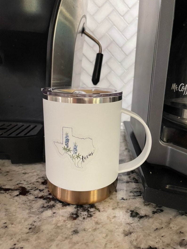 May include: White travel mug with a gold base and a handle. The mug features a sticker of the Texas state outline with bluebonnet flowers and the word "Texas". The mug is next to a coffee machine.