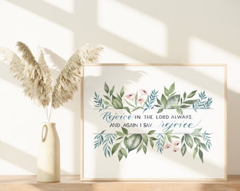 May include: A framed watercolor print with the text "Rejoice in the Lord always, and again I say, rejoice." The artwork features green leaves and pink flowers. A beige vase with pampas grass is to the left.