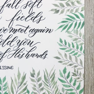May include: A white paper print with elegant black calligraphy text and watercolor botanical illustrations. The text reads "our face, fall soft fields, we meet again, hold you, of His hands, BLESSING". Green leaves and branches frame the edges.