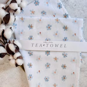 May include: White tea towel with a blue floral pattern and the words "TEA TOWEL 100% COTTON" printed on a white band. The flowers have blue petals and orange centers. A sprig of cotton bolls is in the background.