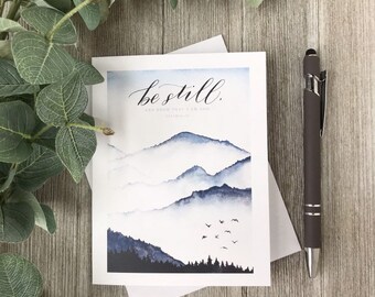 May include: A white card with a watercolor illustration of blue mountains and a flock of birds flying over them. The text "be still. and know that I am God" is written in black calligraphy on the card.