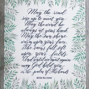 May include: A white print with an Irish blessing in elegant calligraphy. The text is in dark blue, surrounded by a watercolor border of green leaves. The words include "May the road rise up to meet you" and "Irish Blessing."