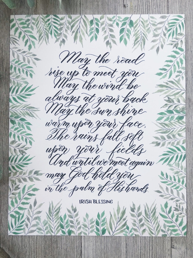 May include: A white print with an Irish blessing in elegant calligraphy. The text is in dark blue, surrounded by a watercolor border of green leaves. The words include "May the road rise up to meet you" and "Irish Blessing."