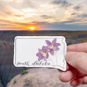 May include: A sticker featuring a purple flower illustration with the text "south dakota" on a white background. The sticker is held in a hand against a blurred background of a sunset over a landscape.