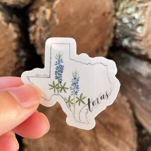 May include: A sticker in the shape of Texas, featuring bluebonnet flowers and the word "Texas" in cursive. The sticker has a white background and a black outline. The bluebonnets are depicted with blue and green watercolor details.