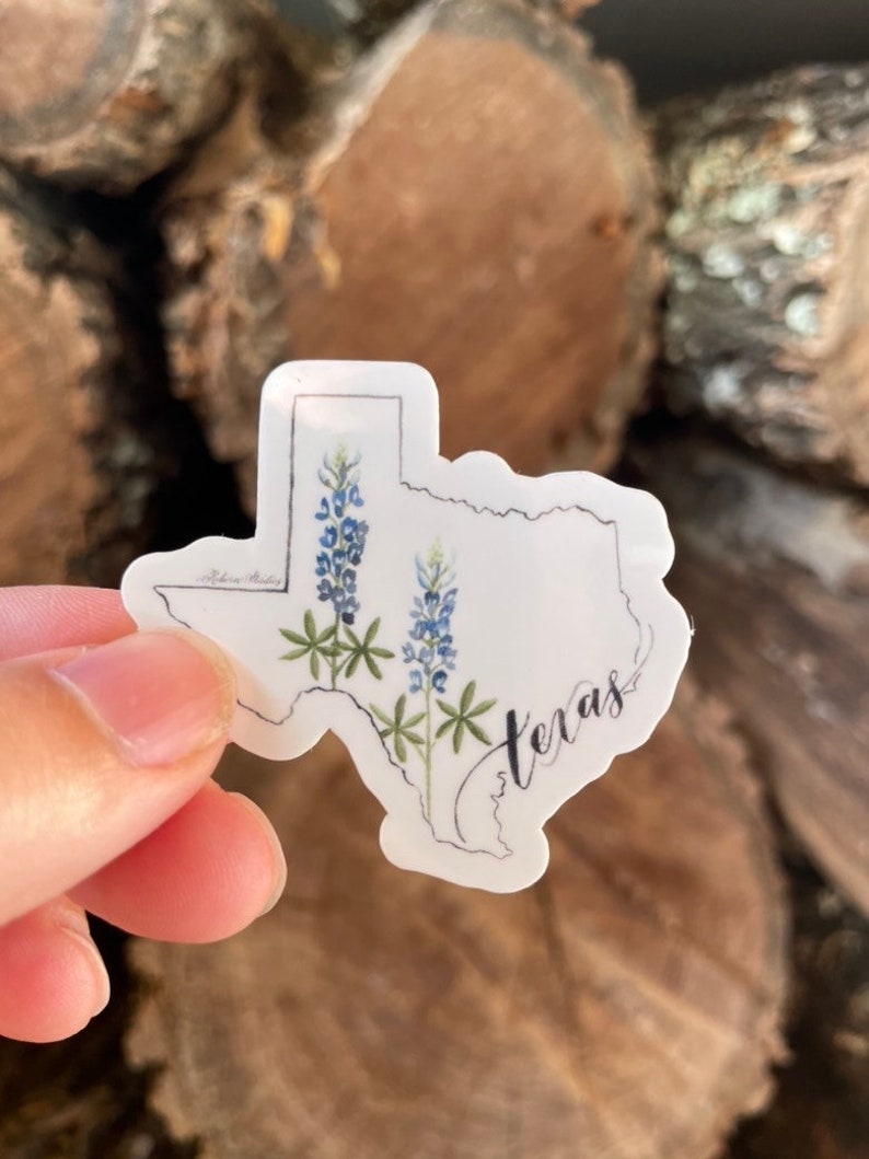 May include: A sticker in the shape of Texas, featuring bluebonnet flowers and the word "Texas" in cursive. The sticker has a white background and a black outline. The bluebonnets are depicted with blue and green watercolor details.