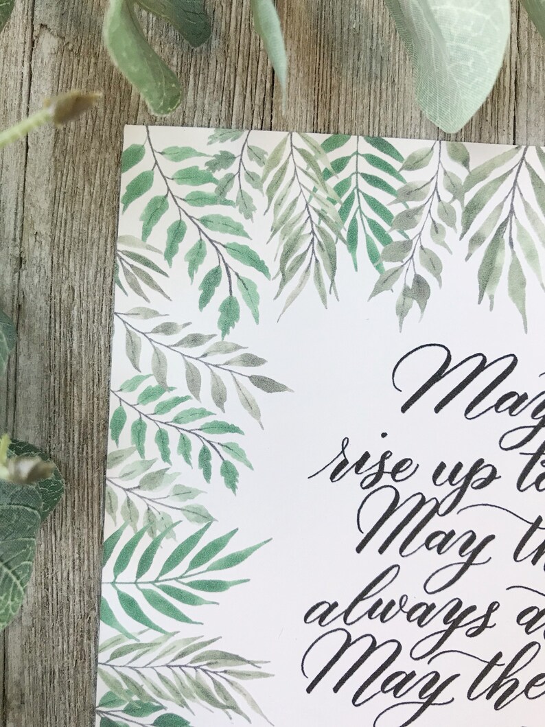 May include: A white card with a watercolor design of green leaves and branches. The card has elegant black calligraphy text that reads "May the..." and continues with additional script. The leaves frame the top and left side of the card.