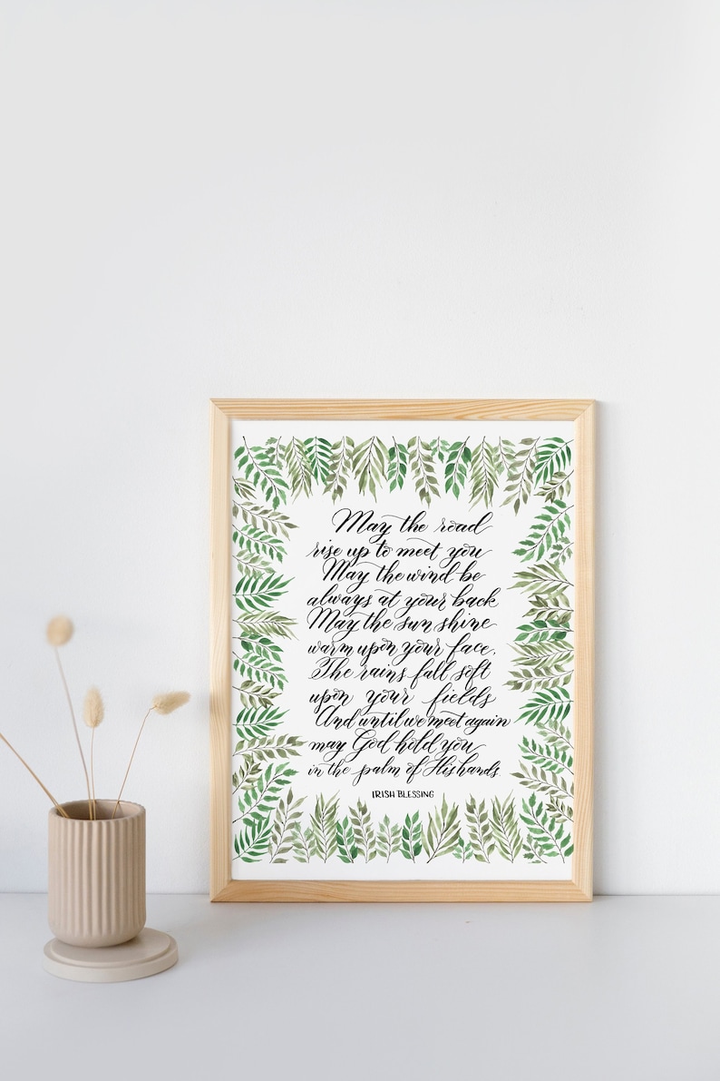 May include: A framed print with an Irish blessing in elegant calligraphy. The text is surrounded by a border of green watercolor leaves. The frame is made of light-colored wood. A small beige vase with dried flowers is on the left.