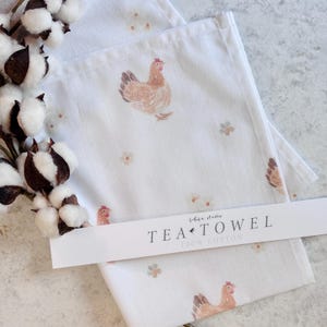 May include: White tea towel with a watercolor illustration of a hen and small flowers. The text "TEA TOWEL 100% COTTON" is printed on a white label. A cotton branch is in the upper left corner.