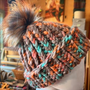 May include: A chunky knit beanie in shades of brown, orange, and teal, topped with a fluffy brown faux fur pom. The hat features a ribbed design and is displayed on a white mannequin head. A cozy winter accessory.