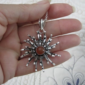 May include: A silver pendant necklace featuring a sunburst design. The pendant has a central, round, orange-brown stone, surrounded by a woven metal pattern and radiating silver bars. The necklace chain is also silver.