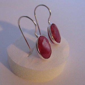May include: A pair of silver-toned drop earrings featuring oval-cut, faceted red gemstones. The gemstones are set in a silver bezel and suspended from curved ear wires. The earrings are displayed on a white surface.