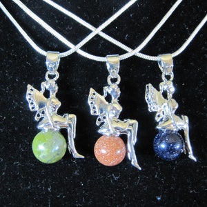 May include: Three silver-toned fairy pendants, each with a seated fairy figure atop a gemstone sphere. The spheres are green, orange, and dark blue, suspended on silver chains. The background is black.