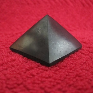 May include: A dark gray pyramid-shaped object with a flat base. The pyramid is made of a solid, possibly stone-like material. The background is a textured, bright red surface.