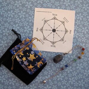 May include: A divination set featuring a pendulum with a faceted gray stone and a chain with colorful beads. Includes a chart with "Yes," "No," and "Maybe" options, and two small pouches, one black and one blue with gold stars and moons.