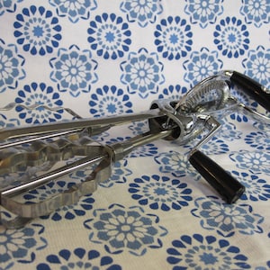 May include: Vintage hand-crank egg beater with a chrome finish and black handles. The beater features a double-gear design and is set against a white and blue floral patterned background. A classic kitchen tool.