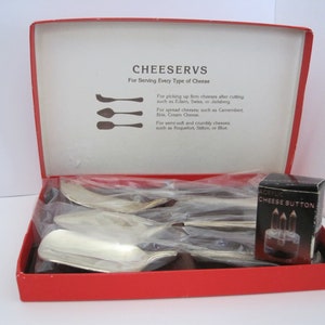 May include: A vintage cheese serving set in a red box. The box is open, revealing silver cheese knives and a small box labeled "ACRYLIC CHEESE BUTTON". The box lid has text that reads "CHEESERVS For Serving Every Type of Cheese".