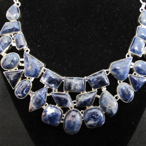 May include: A silver-toned necklace featuring a variety of blue sodalite gemstones in different shapes. The stones are set in a silver metal and arranged in a bib style, creating a striking visual effect. The necklace is displayed against a black background.