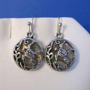 May include: A pair of silver-toned earrings. Each earring features a round, iridescent abalone shell center, framed by a decorative, openwork metal design. The earrings have a simple hook closure.
