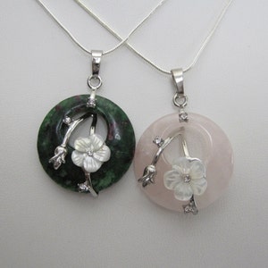 May include: Two pendant necklaces. One features a green stone with a white flower and silver accents. The other has a pink stone with a white flower and silver accents. Both pendants are on silver chains.