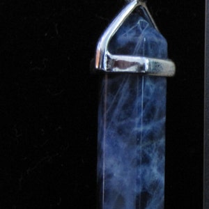 May include: A blue sodalite crystal pendant with a silver-colored metal bail. The crystal is a rectangular shape with a pointed bottom. The blue color has white marbling throughout. The pendant is displayed against a black background.