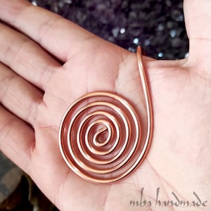 May include: A copper wire pendant shaped into a continuous spiral. The metal has a polished, metallic finish with a warm reddish-brown hue. The spiral design consists of multiple concentric circles that originate from a central point.