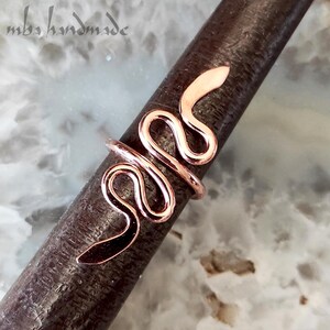 以下が含まれることがあります： 暗い金属棒に巻き付いた銅色のヘビの指輪。リングのデザインは、曲がった体と持ち上げられた頭を持つ様式化されたヘビが特徴です。リングは手作りで、左上に「mbs handmade」の文字があります。