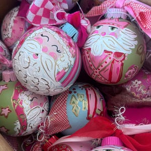 May include: A collection of round Christmas ornaments featuring hand-painted nutcracker and Santa Claus faces in shades of pink, green, and blue. Each ornament is topped with a decorative fabric bow in checkered or solid pink and red patterns.