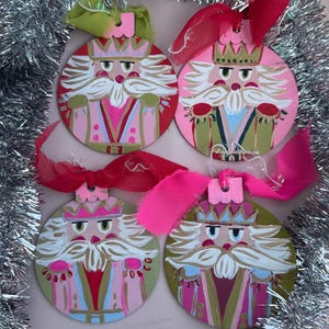 Könnte beinhalten: Vier runde Weihnachtsschmuckstücke mit Nussknacker-Design. Jedes Ornament hat einen rosa oder grünen Hintergrund, ein weißbärtiges Gesicht, eine Krone und eine rot-goldene Uniform. Sie sind mit rosa oder grünen Bändern verziert.
