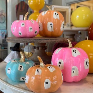 May include: A collection of decorative pumpkins in bright pink, orange, and light blue, each featuring hand-painted white ghost motifs and metallic gold stems. These smooth, rounded gourds are arranged on a wooden pedestal and a white surface.