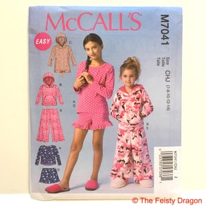 May include: Sewing pattern for girls' pyjamas with a hooded top and trousers. The pattern includes two different styles: one with pink polka dots and one with a camouflage print. The pattern is for sizes 7-8, 10-12, and 14. The pattern is labelled "Easy" and "M7041/CHJ".