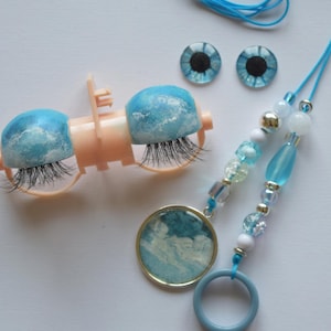 Pode incluir: Uma coleção de materiais de artesanato com um par de cílios postiços azuis e brancos com tema de nuvens, dois enfeites em forma de olho azul e um berloque de contas com um design de nuvem correspondente. O berloque é preso a um anel azul.