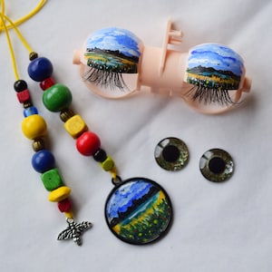 Pode incluir: Um colar de contas de madeira coloridas com um amuleto de abelha e um pingente redondo com uma paisagem pintada. O pingente é preso ao colar com um cordão amarelo. Também estão incluídos dois pares de olhos de boneca de plástico com paisagens pintadas.