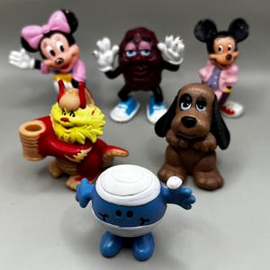 May include: A collection of colorful figurines featuring cartoon characters. The figures include Minnie Mouse in a pink dress, Mickey Mouse in a pink coat, a raisin character, a dog, a dragon, and a blue and white character.