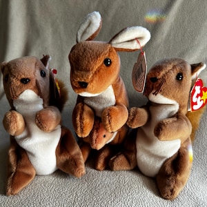 May include: Three plush stuffed animals: two squirrels and a kangaroo. The squirrels are brown with white bellies and fluffy tails. The kangaroo is brown with a joey in its pouch. Each has a tag.