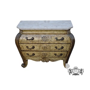 May include: A three-drawer chest with a light beige finish, featuring a rectangular marble top, curved sides, and dark metal hardware. Each drawer is adorned with a central circular floral carving, and the base includes a carved shell detail.