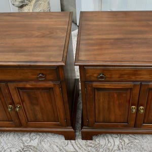 May include: Two matching wooden nightstands with a rich, dark brown finish. Each nightstand features a cabinet with two doors, a drawer, and brass-colored hardware. The tops are rectangular and slightly overhanging.