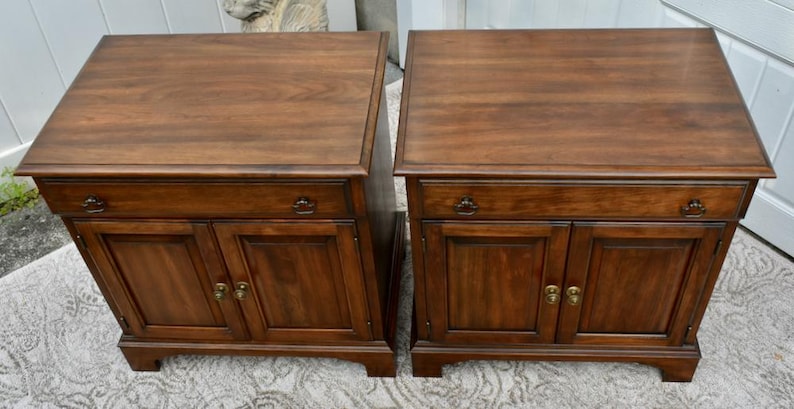 May include: Two matching wooden nightstands with a rich, dark brown finish. Each nightstand features a cabinet with two doors, a drawer, and brass-colored hardware. The tops are rectangular and slightly overhanging.