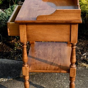 May include: A wooden side table with a warm, medium-brown finish. It features a drawer, a lower shelf, and decorative turned legs. The top has a raised edge on one side. A classic piece of furniture.