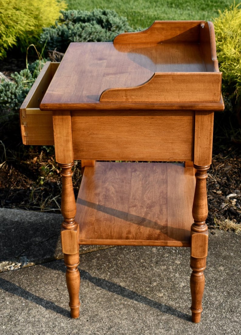 May include: A wooden side table with a warm, medium-brown finish. It features a drawer, a lower shelf, and decorative turned legs. The top has a raised edge on one side. A classic piece of furniture.