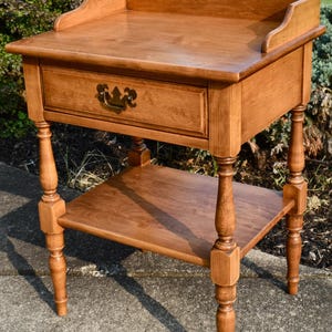 May include: A wooden side table with a drawer and a lower shelf. The table is a warm brown color and has decorative turned legs. The drawer has a brass-colored handle. The top of the table has a raised edge.