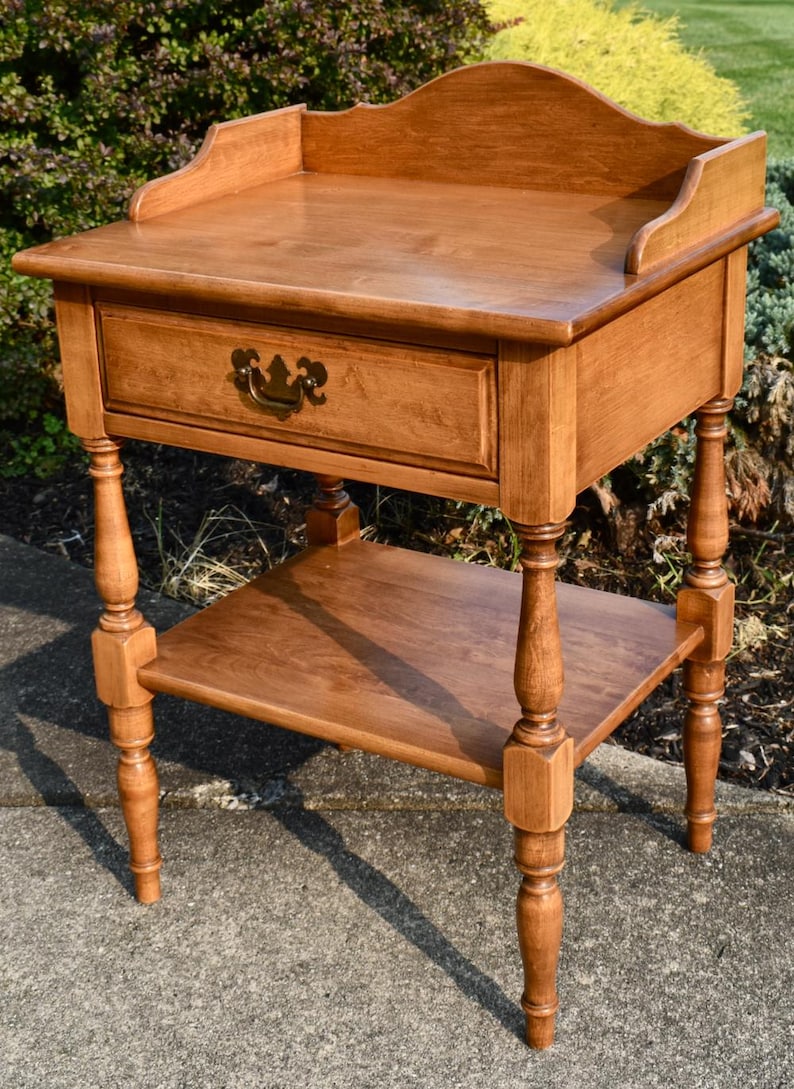 May include: A wooden side table with a drawer and a lower shelf. The table is a warm brown color and has decorative turned legs. The drawer has a brass-colored handle. The top of the table has a raised edge.