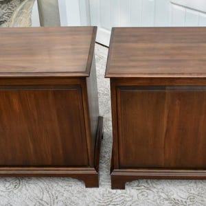 May include: Two matching dark brown wooden side tables. Each table has a rectangular top, a paneled front, and a base with short legs. The wood grain is visible, and the tables appear to be in good condition.