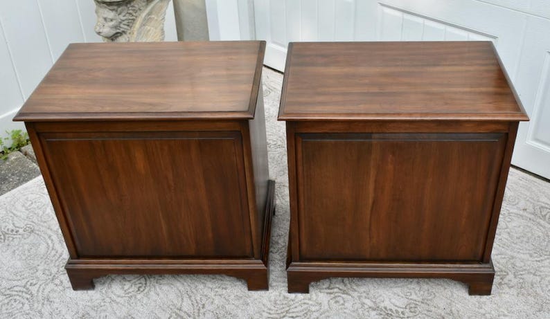 May include: Two matching dark brown wooden side tables. Each table has a rectangular top, a paneled front, and a base with short legs. The wood grain is visible, and the tables appear to be in good condition.